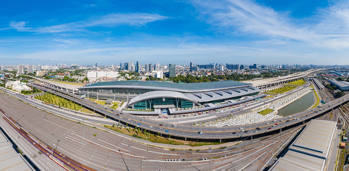 Aerial view of Krung Thep Aphiwat Central Terminal on a sunny day, showing its architectural details, surrounded by Bangkok's towering buildings against a clear blue sky.