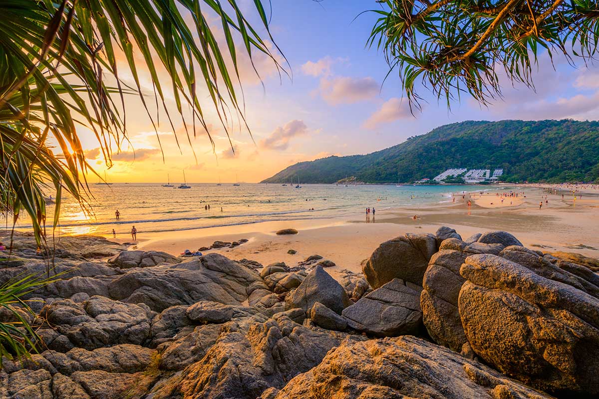 Sunset at Nai Harn Beach, Phuket, with an orange sky, swimmers, people on the sandy beach, sailboats in the sea, and rocks along the coastline.