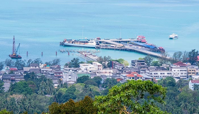 Panoramic shot from a mountain in Koh Samui on March 23, 2023, highlighting the serene Nathon Bay, vibrant Nathon Beach, and structured Nathon Pier with a backdrop of the calm sea and lush coconut tree forest.