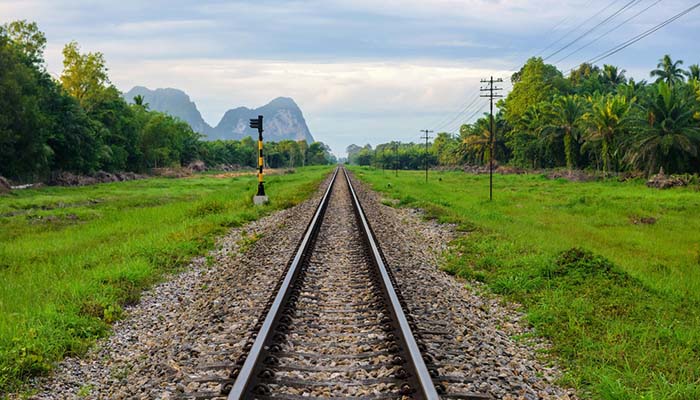 Morning view of a straight railway with palm tree farm and mountain backdrop in Surat Thani, Thailand.